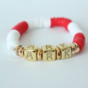 May include: A red and white beaded bracelet with gold letter beads spelling out the name "ARK".