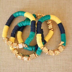 Op de afbeelding: Drie kleurrijke armbanden met gouden kralen. De armbanden zijn gemaakt van gele, groene en blauwe kralen. De gouden kralen zijn vierkant en hebben letters erop.