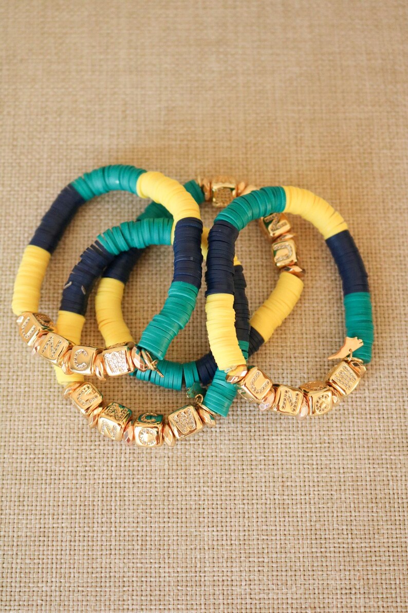 Op de afbeelding: Drie kleurrijke armbanden met gouden kralen. De armbanden zijn gemaakt van gele, groene en blauwe kralen. De gouden kralen zijn vierkant en hebben letters erop.