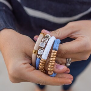 May include: A stack of four bracelets with different textures and colors. The bracelets are made of wood, metal, and beads. The bracelets are in shades of blue, white, and gold.