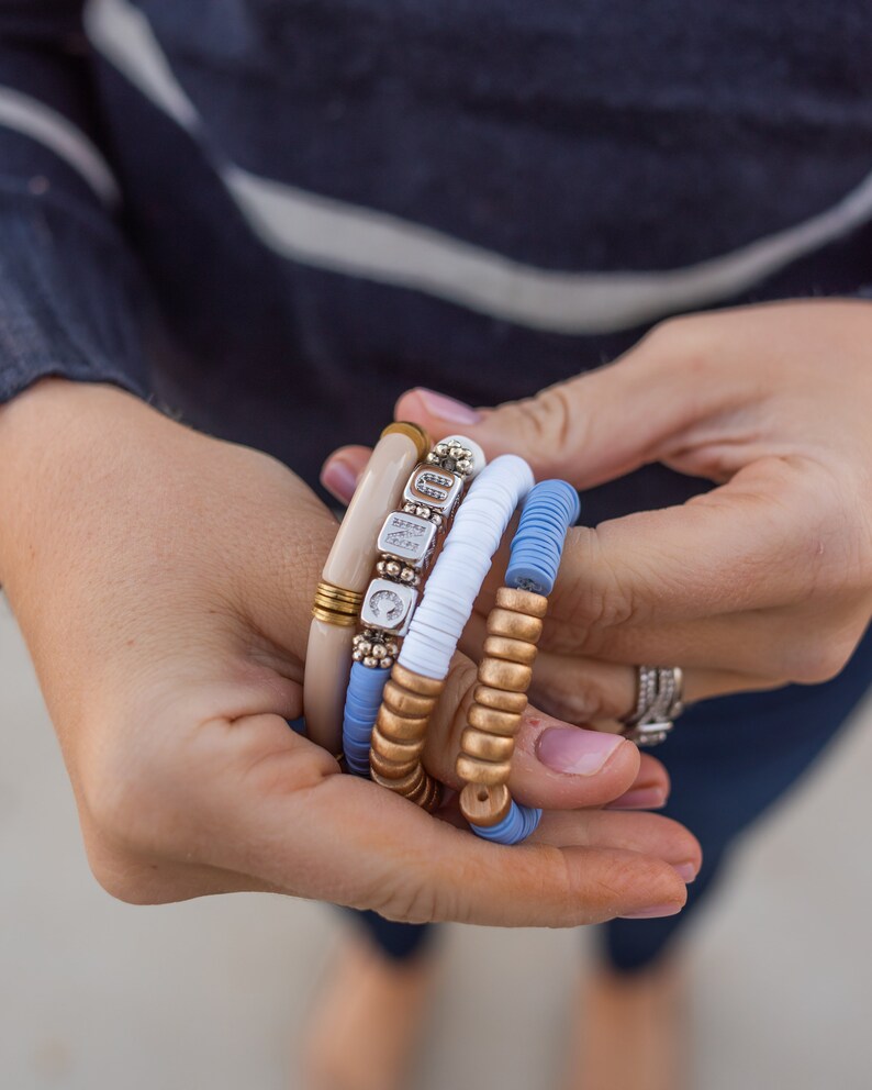 May include: A stack of four bracelets with different textures and colors. The bracelets are made of wood, metal, and beads. The bracelets are in shades of blue, white, and gold.
