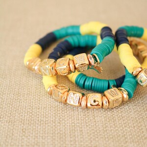 Op de afbeelding: Drie armbanden met gele, turquoise en donkerblauwe kralen. De kralen zijn geregen aan een elastisch koord en hebben gouden letterbedels die het woord "UNC" spellen.