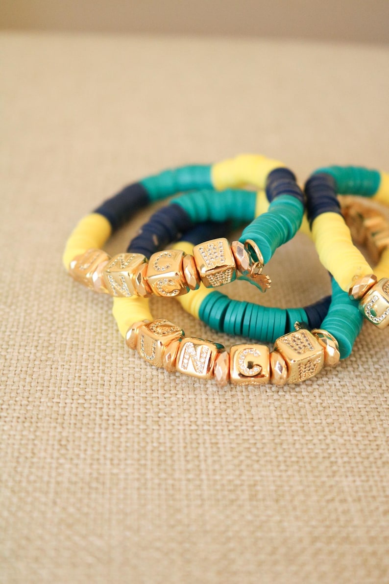 Op de afbeelding: Drie armbanden met gele, turquoise en donkerblauwe kralen. De kralen zijn geregen aan een elastisch koord en hebben gouden letterbedels die het woord "UNC" spellen.