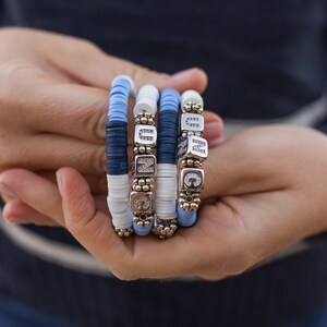 May include: Four stacked bracelets with blue and white beads and silver charms with letters and rhinestones.