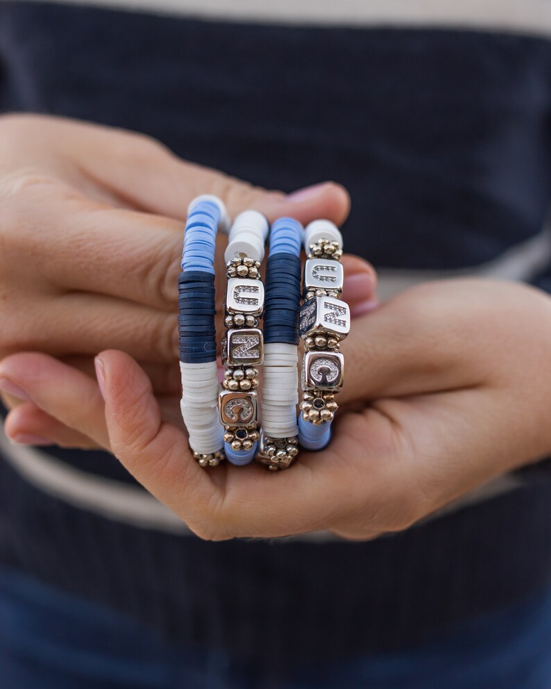 May include: Four stacked bracelets with blue and white beads and silver charms with letters and rhinestones.