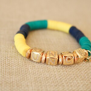 Op de afbeelding: Een kleurrijke armband met gele, blauwe en groene kralen. De armband heeft gouden bedelarmbanden met de letters "UNC" erop.