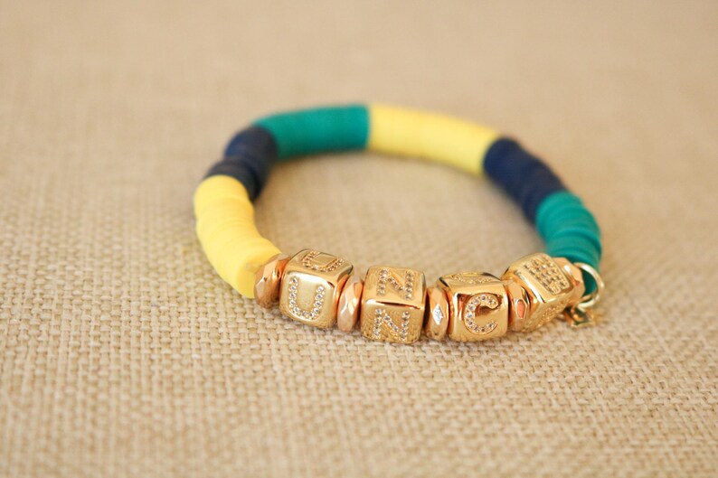 Op de afbeelding: Een kleurrijke armband met gele, blauwe en groene kralen. De armband heeft gouden bedelarmbanden met de letters "UNC" erop.