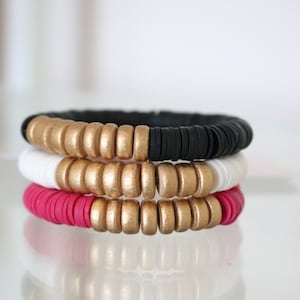 May include: A set of three bracelets with gold, white, and pink beads. The bracelets are stacked on top of each other.