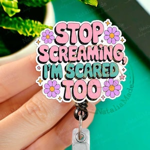 May include: A pink and green badge reel with the text "STOP SCREAMING, I'M SCARED TOO" surrounded by pink and purple flowers. The badge reel is attached to a silver retractable clip.