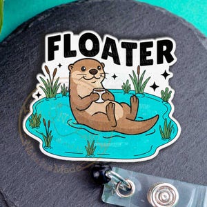 May include: A badge reel with a cartoon otter relaxing in a blue pond, holding a drink, with the word "FLOATER" above. The reel has a clear plastic holder and a black retractable cord.