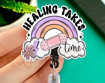 May include: A pink and purple rainbow with a white bandage and a lightning bolt. The rainbow is surrounded by clouds with the text "Healing Takes Time". The rainbow is attached to a black and silver retractable badge reel.
