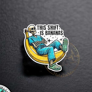 May include: A 1.25 inch acrylic pin featuring a skeleton wearing a blue scrubs outfit and sitting on a yellow banana. The skeleton is wearing green sandals and has a stethoscope around its neck. The text "THIS SHIFT IS BANANAS" is above the skeleton.