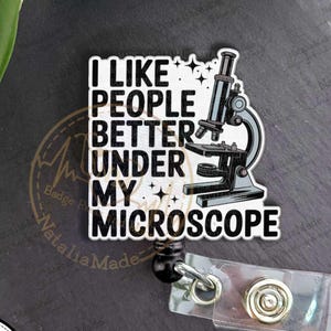 May include: A white and black badge reel with the text "I LIKE PEOPLE BETTER UNDER MY MICROSCOPE" and a microscope graphic. The badge reel has a black retractable cord and a clear plastic ID holder.