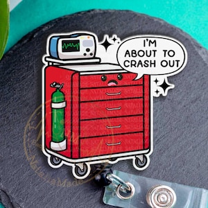 May include: A red medical cart badge reel with a cartoon design. The cart has drawers, an oxygen tank, and a monitor. Text reads "I'M ABOUT TO CRASH OUT". The badge reel has a clear plastic strap and a metal clip.