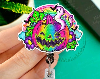 May include: A rainbow glitter pumpkin with a spooky face, surrounded by clouds, bats, and flowers. The pumpkin is attached to a black retractable badge reel with a silver clip.