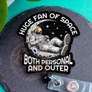 May include: A black and white badge reel with the text "HUGE FAN OF SPACE" and "BOTH PERSONAL AND OUTER". The badge reel features an astronaut relaxing on the moon. The badge reel is on a black slate coaster.