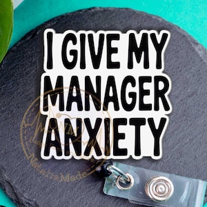 May include: A white badge reel featuring the black text I GIVE MY MANAGER ANXIETY in a bold, sans-serif font. The badge reel includes a clear plastic strap with a metal snap button and a retractable cord mechanism for holding identification cards.