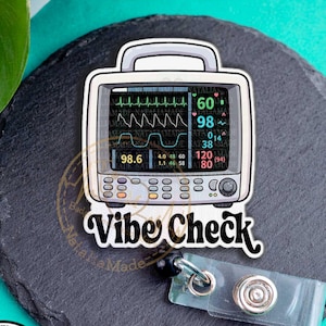 May include: A white and gray badge reel featuring a medical monitor design with the text "Vibe Check." The monitor displays vital signs. Includes a clear plastic badge holder and a black retractable cord.