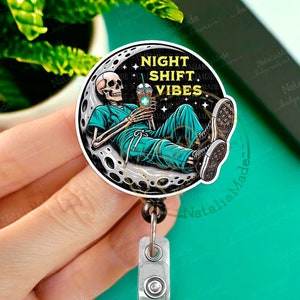 May include: A black and white cartoon skeleton wearing a blue scrubs uniform and holding a cup of coffee. The skeleton is sitting on a crescent moon with the text "Night Shift Vibes" above it. The image is on a round badge reel.