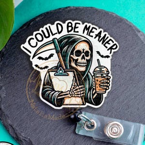 May include: A badge reel with a cartoon grim reaper holding a clipboard and a drink, with the text "I COULD BE MEANER". The reel is on a black slate-like surface with a badge reel clip.