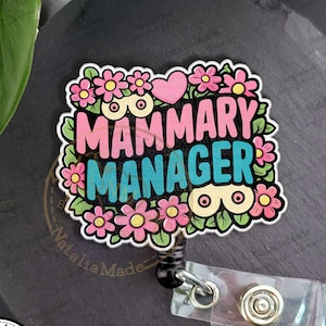May include: A badge reel with the text "Mammary Manager" in pink and teal, surrounded by pink flowers and green leaves. A pink heart is above the word "Manager". The reel has a clear plastic strap and a black retractable cord.