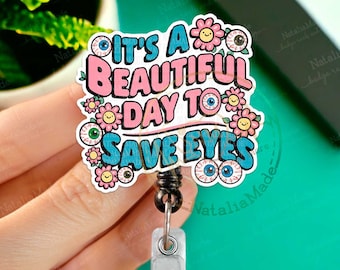 May include: A pink and blue badge reel with the text "It's a beautiful day to save eyes". The text is surrounded by colorful flowers and cartoon eyes. The reel is attached to a black retractable cord and a silver clip.