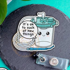 May include: A badge reel featuring a cartoon illustration of a medical suction canister with the text "It's ok to suck at new things." The reel has a clear plastic clip and a black retractable cord.