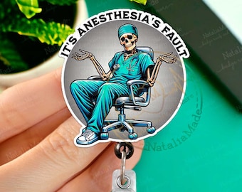 Könnte beinhalten: Ein runder Badge-Halter mit einem Skelett, das einen blauen OP-Kittel und ein Stethoskop trägt. Der Badge-Halter trägt den Text "Es ist die Schuld der Anästhesie" in einem weißen Kreis auf grauem Hintergrund.
