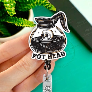 May include: A black and white coffee pot with a skull inside, with the text "POT HEAD" on the bottom. The coffee pot is attached to a black retractable badge reel with a silver clip.