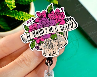 May include: A black and white skull with pink and purple flowers and a banner that reads "BE AFRAID & DO IT ANYWAY". The skull is attached to a black and silver retractable badge reel.