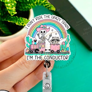 May include: A colourful badge reel featuring a skeleton wearing a cowboy hat and riding a train with a raccoon. The text reads "I don't ride the crazy train I'm the conductor".