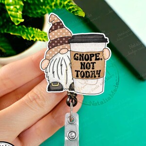 May include: A brown and white gnome holding a coffee cup with the text "GNOPE, NOT TODAY". The gnome is wearing a brown hat with white polka dots. The coffee cup is brown with a white lid. The gnome and coffee cup are attached to a black retractable badge reel with a silver clip.