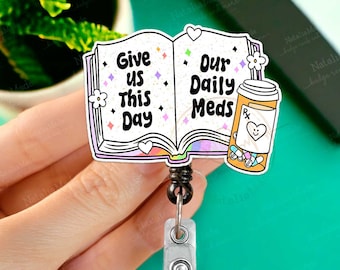 May include: A colorful book-shaped badge reel with the text "Give us this day our daily meds" and a pill bottle with a heart on it. The reel is attached to a black retractable cord and a silver clip.