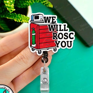 May include: A red medical trolley with a black retractable badge reel. The trolley has a green oxygen tank and a medical monitor. The text on the badge reel says "We Will ROSC You".