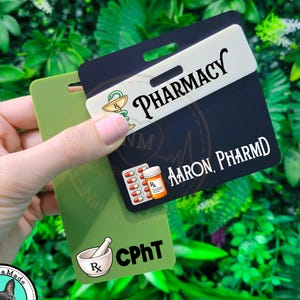 May include: Two rectangular plastic badge buddies. The top black badge features the text PHARMACY, a medical symbol, a pill blister pack, an orange pill bottle, and the name AARON, PHARMD. The bottom olive green badge displays a mortar and pestle and CPhT.