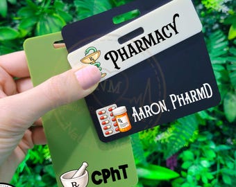 May include: Two rectangular plastic badge buddies. The top black badge features the text PHARMACY, a medical symbol, a pill blister pack, an orange pill bottle, and the name AARON, PHARMD. The bottom olive green badge displays a mortar and pestle and CPhT.