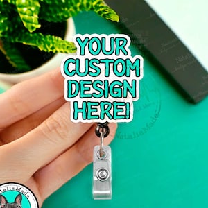 May include: A teal blue badge reel with a white circle and the text "YOUR CUSTOM DESIGN HERE!" in black lettering. The reel is attached to a black retractable cord and a silver clip.