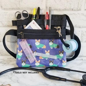May include: Black nylon medical organizer bag with a clear front panel featuring a pattern of corgis wearing face masks and sweaters. The bag has multiple pockets, a zipper closure, and a shoulder strap. Includes scissors, pens, and tape.