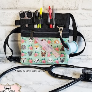 May include: Black medical utility belt with a teal front pocket featuring a dog and floral pattern. The belt has multiple pockets and loops for tools, including scissors, pens, and a small spool. A stethoscope is in the foreground.