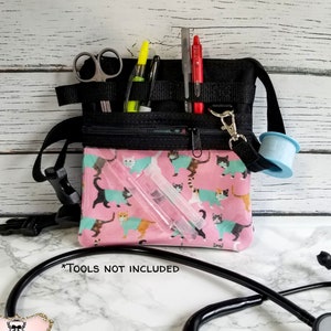 May include: Black and pink medical fanny pack with a cat print. The pack has multiple pockets and compartments for medical tools. The image includes scissors, pens, and tape. The text on the image says "TOOLS NOT INCLUDED".