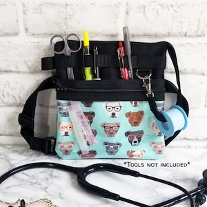May include: Black and teal medical fanny pack with a dog-in-glasses pattern. The pack has multiple pockets and loops for tools, including scissors, pens, and tape. A stethoscope is in the foreground. The text "*TOOLS NOT INCLUDED*" is visible.