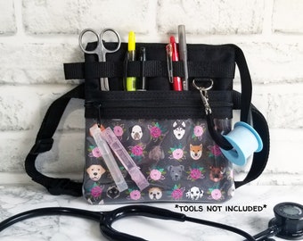 May include: Black medical fanny pack with a clear front pocket featuring a dog and floral pattern. The pack includes a black strap, scissors, pens, and tape. A black stethoscope rests in front of the bag. The text "*TOOLS NOT INCLUDED*" is visible.