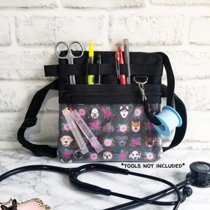 May include: Black medical fanny pack with a clear front pocket featuring a dog and floral pattern. The pack includes a black strap, scissors, pens, and tape. A black stethoscope rests in front of the bag. The text "*TOOLS NOT INCLUDED*" is visible.
