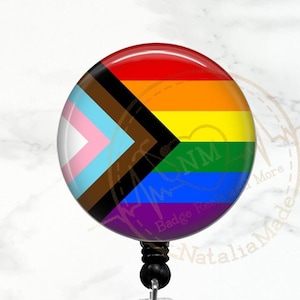 May include: A rainbow pride flag with a black and brown stripe, a light blue stripe, a light pink stripe, a dark blue stripe, a purple stripe, a light green stripe, a yellow stripe, and a red stripe. The flag is on a retractable badge reel with a clear plastic clip.