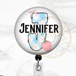 May include: A round badge reel featuring the name "Jennifer". The letter "J" is in a blue and white plaid design, with a pink strawberry and bow. The badge reel has a clear plastic clip and a black retractable cord.