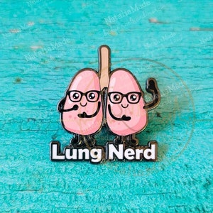 May include: A 1.25 inch enamel pin featuring a cartoon illustration of two pink lungs wearing glasses and smiling. The lungs are connected by a black line and the text "Lung Nerd" is written below them.