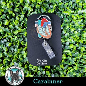 May include: A badge reel with a carabiner clip, featuring a colorful anatomical heart design. The heart illustration includes labels for the aorta, atrium, and ventricles. The text "No Clip On Back" is visible. The bottom of the reel reads "Carabiner".