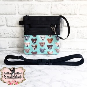 May include: Black and teal dog treat bag with a matching adjustable strap. The bag features a teal background with a pattern of dogs wearing glasses. The bag has a black zipper and a black strap with a plastic buckle.