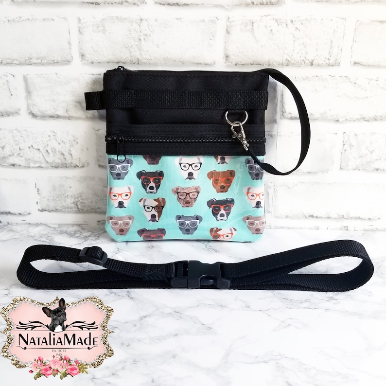 May include: Black and teal dog treat bag with a matching adjustable strap. The bag features a teal background with a pattern of dogs wearing glasses. The bag has a black zipper and a black strap with a plastic buckle.
