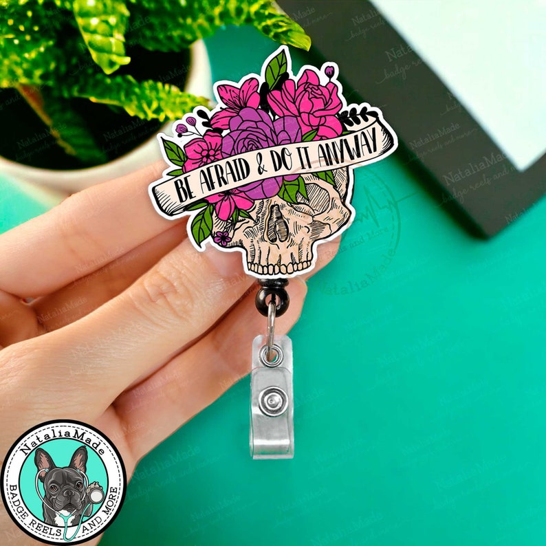 May include: A black and white skull with pink and purple flowers and a banner that reads "BE AFRAID & DO IT ANYWAY". The skull is attached to a black and silver retractable badge reel.